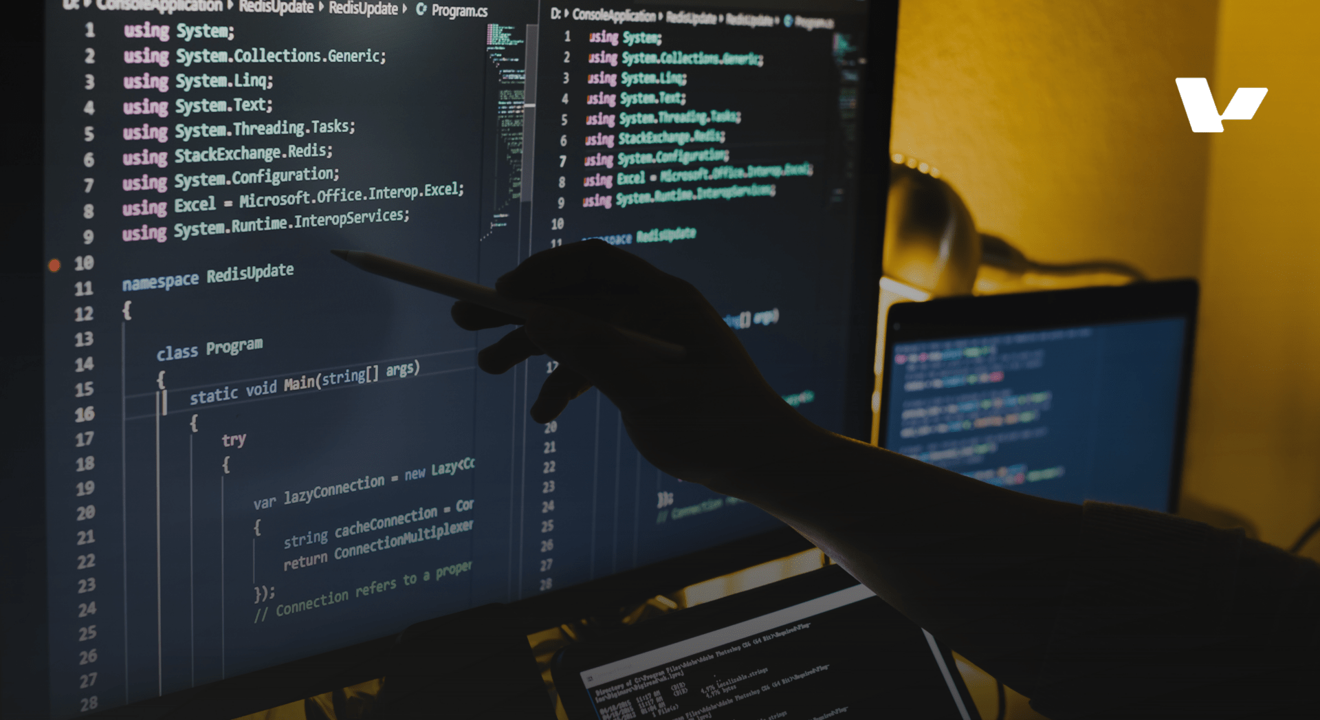 A programmer using a stylus to point at lines of C# code on a dark computer monitor, with a laptop and warm ambient lighting in the background.